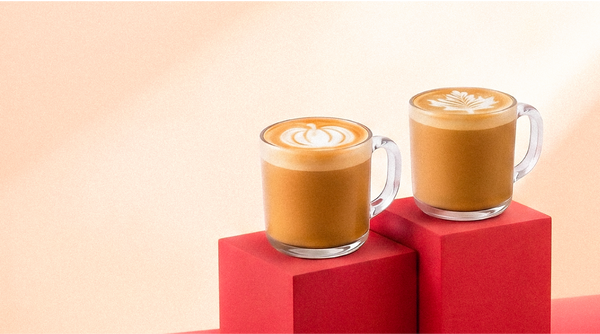 Two clear glass mugs of maple pecan and pumpkin spice lattes, topped with latte art featuring a maple leaf and pumpkin, displayed on red geometric blocks against a soft peach background.