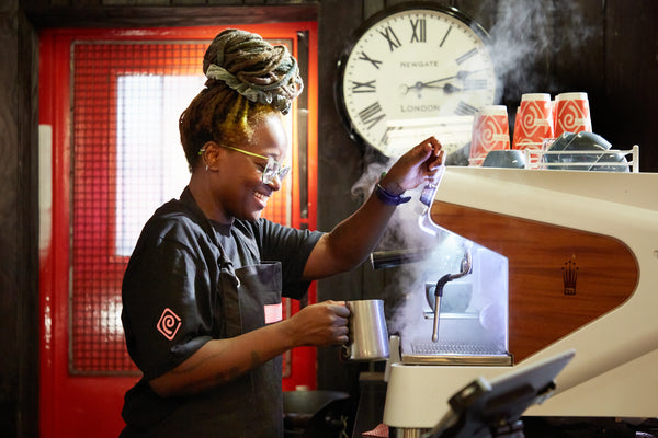 Person operating a coffee machine with a red wall and clock in the background