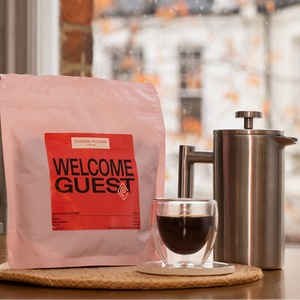 Coffee bag labeled 'Welcome Guest' with a glass of coffee and French press on a wooden surface.